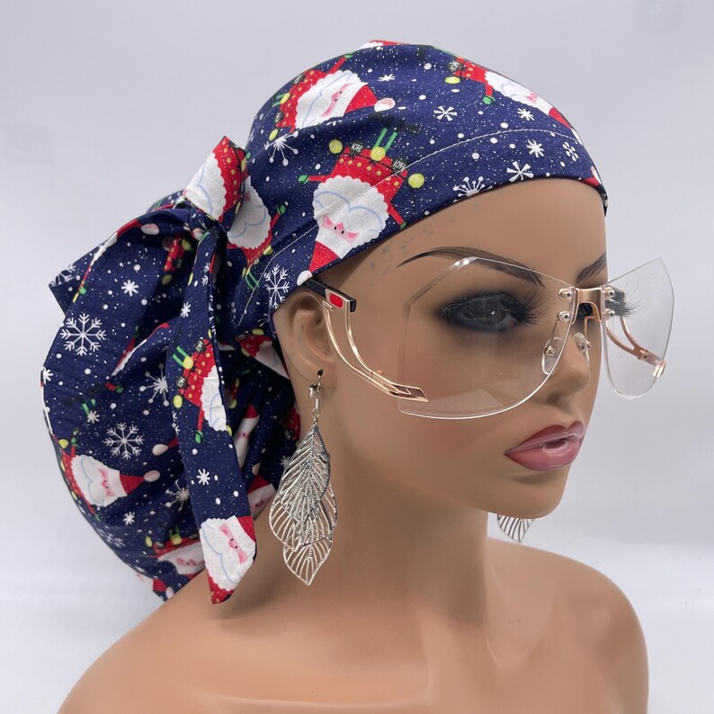 May include: A navy blue scrub cap with a Christmas Santa Claus print, tied in a bow. The cap is paired with gold-rimmed, clear-lens glasses and silver leaf earrings. The cap features a pattern of Santa Claus figures and snowflakes.