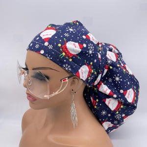 May include: A navy blue bouffant scrub cap featuring a festive Santa Claus and snowflake pattern. The cap is made of fabric and is designed to cover the hair. The Santa Claus figures are red and white. The background is navy blue.