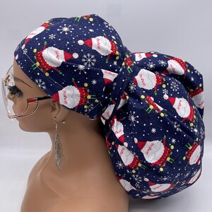 May include: A navy blue scrub cap featuring a festive Santa Claus and snowflake pattern. The cap is made of fabric and is designed to cover the hair. The Santa Claus figures are in red and white, with green accents. The background is navy blue.