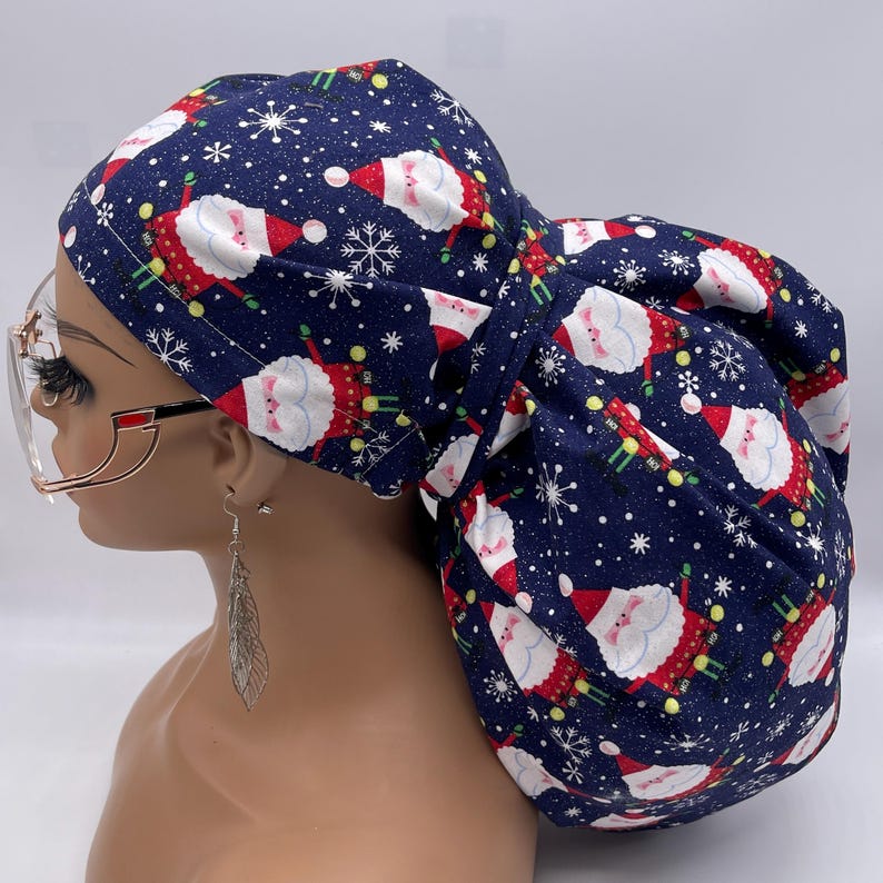 May include: A navy blue scrub cap featuring a festive Santa Claus and snowflake pattern. The cap is made of fabric and is designed to cover the hair. The Santa Claus figures are in red and white, with green accents. The background is navy blue.