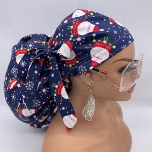 May include: A navy blue scrub cap featuring a festive Santa Claus print with snowflakes. The cap is tied in a bow at the back. The Santa Claus design is in red, white, and green. The cap is designed for medical professionals.