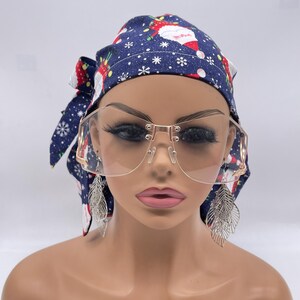 May include: A mannequin head is adorned with a navy blue headscarf patterned with Santa Claus and snowflakes. The mannequin wears large, rimless, gold-framed sunglasses and silver feather earrings. The overall theme is festive and stylish.