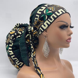 May include: A black, green, and yellow patterned fabric head wrap with a large bun on top. The wrap is tied at the back with long ties. The wrap is worn by a mannequin with silver leaf earrings.