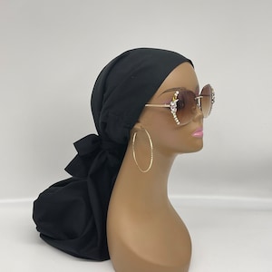 May include: A black satin head wrap with a bow tied at the back. The wrap is worn on a mannequin with brown skin and large gold hoop earrings. The mannequin is wearing large, gold-rimmed sunglasses with rhinestones.