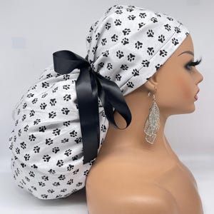 May include: A white and black paw print surgical scrub cap with a black satin ribbon. The cap is tied in a bow at the back. The cap is designed to cover the hair during medical procedures.
