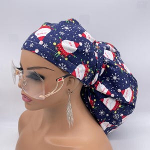May include: A navy blue bouffant scrub cap with a Santa Claus and snowflake pattern. The cap is paired with clear, oversized rimless glasses with gold frames and a red accent. Silver feather earrings complete the look.