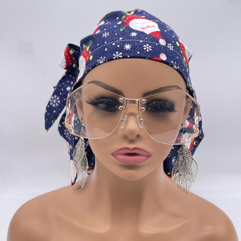 May include: A mannequin head is adorned with a navy blue Christmas-themed headscarf and large, clear-lens sunglasses with a gold frame. Silver leaf earrings dangle from the ears. The headscarf features a pattern of Santa Claus, snowflakes, and red accents.
