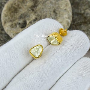 May include: A pair of gold stud earrings featuring a light green, triangular gemstone. The earrings are set in a gold bezel. The image includes the text "Fine Jewellerys".