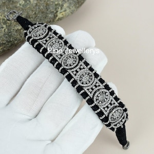 Puede incluir: Pulsera negra con adornos circulares plateados. La pulsera tiene una banda negra tejida con un patrón de cuadrícula. Los adornos circulares están incrustados con pequeñas piedras transparentes. El texto "Pine Jewellerys" es visible.