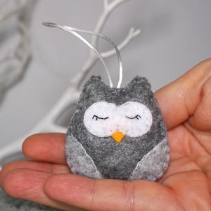 Könnte beinhalten: Handgefertigter Eulen-Anhänger aus Filz in Grau, Weiß und Gelb. Die Eule hat geschlossene Augen, einen kleinen orangen Schnabel und eine silberne Aufhängeschlaufe. Als dekoratives Element konzipiert.