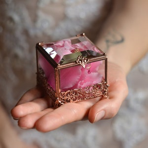 May include: A small, square, rose gold ring box with a clear glass top and a pink flower inside. The box is ornate with a delicate, filigree design.