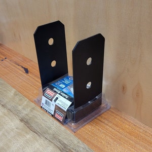 May include: Black metal outdoor post brackets in a clear plastic package with a red and white label that says "Outdoor Accents" and "Simpson Strong-Tie".