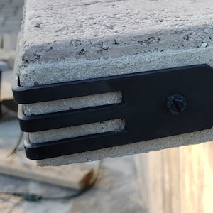 May include: Close-up of a black metal corner bracket attached to a concrete surface with a screw. The bracket has three horizontal slots.