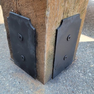 Può includere: Due staffe in metallo nero con bordi decorativi sono fissate a un palo di legno. Le staffe hanno due fori per viti ciascuna.