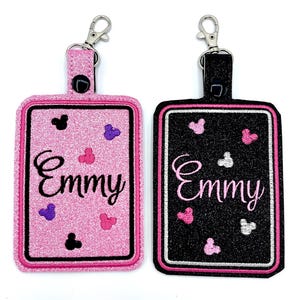 May include: Two pink and black luggage tags with a silver clip. The tags have a black Mickey Mouse head and the name "Emmy" embroidered on them.