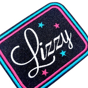 Könnte beinhalten: Ein schwarzer Bügel-Patch mit dem Namen "Lizzy" in weißer Kursivschrift. Der Patch hat einen doppelten rosa und blauen Rand und vier weiße Sterne.