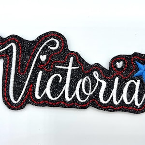 Puede incluir: Parche de planchado de brillo negro con letras blancas en cursiva que dice "Victoria" con dos corazones rojos y una estrella azul.