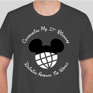 Op de afbeelding: Een donkergrijs T-shirt met een witte afbeelding van Mickey Mouse oren met een wereldbol erin. De tekst "Celebrating My 21st Birthday Drinking Around The World" staat rond de afbeelding.