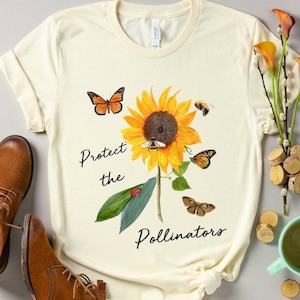 May include: Cream-colored t-shirt featuring a watercolor sunflower with butterflies, bees, and the text "Protect the Pollinators." The shirt is paired with brown leather boots. A cup of coffee and decorative wood slices are also in the image.
