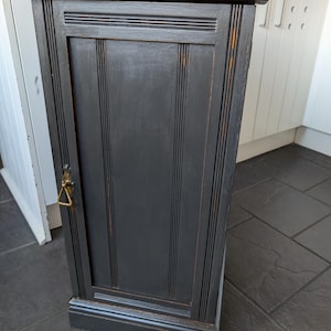 Puede incluir: Un armario de madera negro con una sola puerta y una manija de latón. El armario tiene una tapa de madera y una moldura decorativa alrededor de la puerta.