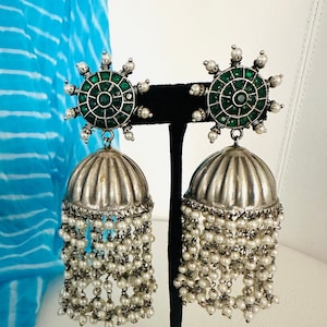 May include: Silver and green dangle earrings with a round, green stone center surrounded by smaller stones. The earrings have a silver bell-shaped charm and a long, beaded fringe.