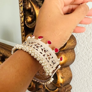 May include: A silver bangle bracelet with intricate floral designs and red gemstone accents. The bracelet is adorned with a row of white pearls.