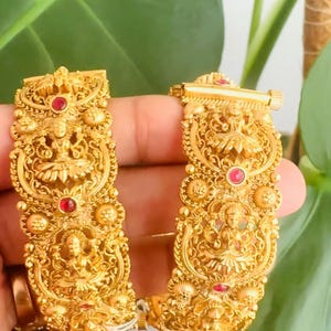 May include: A pair of gold bangles with intricate floral designs and red gemstones. The bangles are made of a traditional Indian style.
