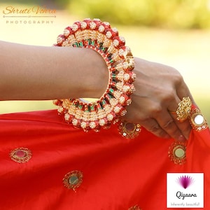 May include: A red, gold, and green bangle with white pearls and a floral design. The bangle is worn on a person's wrist. The person is wearing a red dress with gold embroidery.