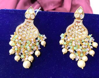 May include: Gold-toned chandelier earrings with a teardrop top and a floral filigree design. The earrings feature clear stones, small green and pink beads, and large cream-colored faux pearls. The earrings are displayed on a blue surface.