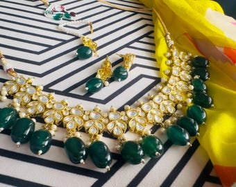 May include: A gold necklace with a large, ornate pendant featuring white stones and green beads. The necklace is paired with matching earrings with green stones.