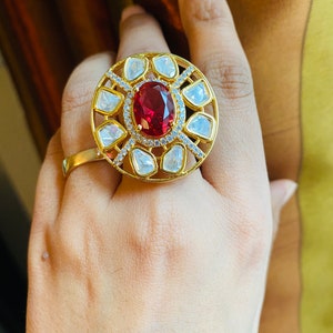 May include: A gold ring with a large red gemstone surrounded by smaller white gemstones. The ring is set in a circular design with a delicate filigree pattern.