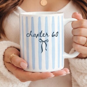 May include: White ceramic mug with blue vertical stripes and the text "chapter 60" with a bow illustration. The mug has a white handle and is held by a person wearing a white sweater. The mug is a perfect gift for a milestone birthday.