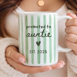 May include: White ceramic mug with green vertical stripes and the text "promoted to auntie" in black, with a green heart and "EST. 2026". The mug has a white handle and is held by a person.