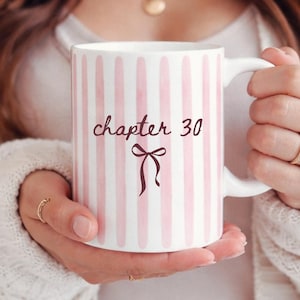 May include: White ceramic mug with pink vertical stripes. The mug features the text "chapter 30" in a script font, along with a bow illustration. The mug is held by a person wearing a white sweater.