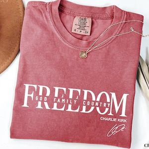 Puede incluir: Camiseta de color carmes&iacute; con la palabra "FREEDOM" impresa en blanco, junto con "GOD FAMILY COUNTRY" y "CHARLIE KIRK". Un collar dorado con un colgante descansa sobre la camiseta. La camiseta tiene una etiqueta Comfort Colors.
