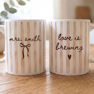 Peut inclure: Deux mugs en céramique blanche à rayures beiges verticales. L'un porte l'inscription "mrs. smith" avec un nœud, l'autre "love is brewing" avec un cœur. Les deux mugs ont une anse.