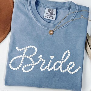 May include: A blue jean colored t-shirt with the word "Bride" spelled out in white pearl-like beads. A gold necklace with a pendant rests on the shirt. The shirt has a Comfort Colors tag.