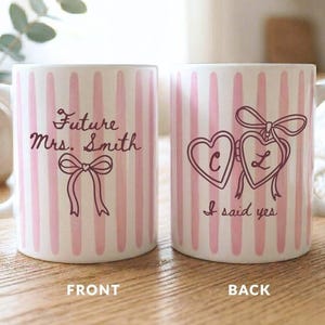 May include: Two white ceramic mugs with vertical pink stripes. The front mug features the text Future Mrs. Smith and a bow illustration. The back mug displays two heart-shaped lockets with the letters C and L, a bow, and the text I said yes.