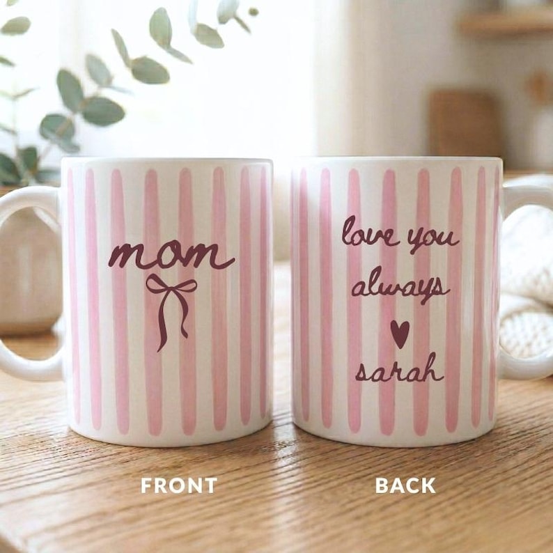 May include: Two white ceramic mugs with pink vertical stripes. One mug says "mom" with a bow graphic, the other says "love you always" with a heart and "sarah".