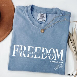 Puede incluir: Una camiseta azul Comfort Colors con la palabra "FREEDOM" impresa en blanco, junto con "GOD FAMILY COUNTRY" y la firma de Charlie Kirk. Un collar dorado con un colgante descansa sobre la camiseta.