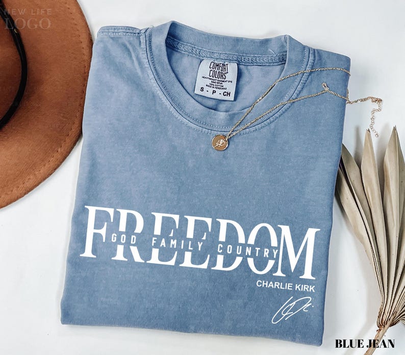 Puede incluir: Una camiseta azul Comfort Colors con la palabra "FREEDOM" impresa en blanco, junto con "GOD FAMILY COUNTRY" y la firma de Charlie Kirk. Un collar dorado con un colgante descansa sobre la camiseta.