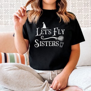 May include: Black t-shirt with white text that reads "Let's Fly Sisters" with a witch's hat and broom graphic.