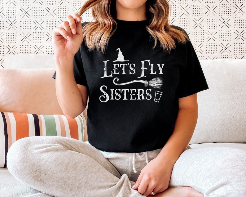May include: Black t-shirt with white text that reads "Let's Fly Sisters" with a witch's hat and broom graphic.