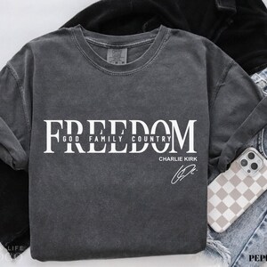 Puede incluir: Camiseta de manga larga gris oscuro con la palabra "FREEDOM" en letras blancas grandes. Debajo, las palabras "GOD FAMILY COUNTRY" y el nombre "CHARLIE KIRK" con una firma. Se ve un tel&eacute;fono con una funda a cuadros.