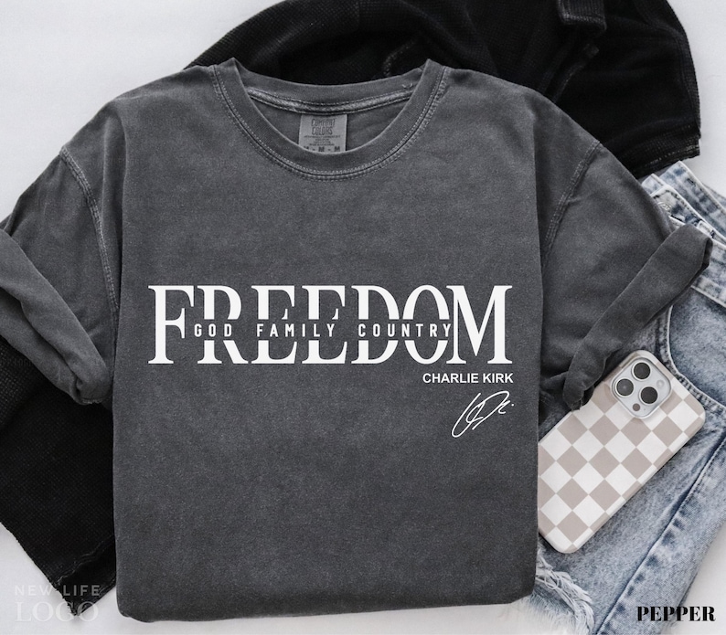 Puede incluir: Camiseta de manga larga gris oscuro con la palabra "FREEDOM" en letras blancas grandes. Debajo, las palabras "GOD FAMILY COUNTRY" y el nombre "CHARLIE KIRK" con una firma. Se ve un tel&eacute;fono con una funda a cuadros.