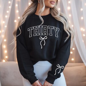 May include: Black sweatshirt with white text that says "THIRTY" with a white bow on each side of the word. The sweatshirt has long sleeves and a crew neck.