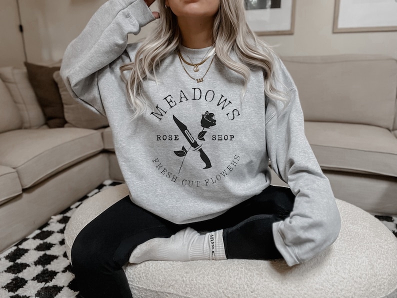 May include: A gray sweatshirt with a black graphic design featuring a rose and a knife. The text on the sweatshirt reads "Meadows Rose Shop Fresh Cut Flowers".