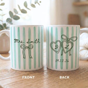 K&ouml;nnte beinhalten: Wei&szlig;e Keramikbecher mit einem mintgr&uuml;nen Streifenmuster. Ein Becher zeigt "Mrs. Smith" und eine Schleifen-Grafik. Der andere Becher hat zwei herzf&ouml;rmige Medaillons mit den Buchstaben "D" und "L" und dem Datum "09.12.26".