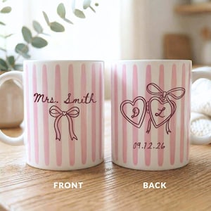 K&ouml;nnte beinhalten: Zwei wei&szlig;e Keramikbecher mit rosa vertikalen Streifen. Ein Becher hat "Mrs. Smith" und ein Schleifendesign. Der andere Becher hat zwei herzf&ouml;rmige Medaillons mit den Buchstaben "D" und "L" und dem Datum "09.12.26".