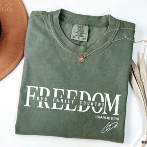 Puede incluir: Una camiseta verde musgo con la palabra "FREEDOM" impresa en blanco, junto con "GOD FAMILY COUNTRY" y la firma "CHARLIE KIRK". Un collar dorado con un colgante descansa sobre la camiseta. Un sombrero marr&oacute;n y una planta seca est&aacute;n en el fondo.